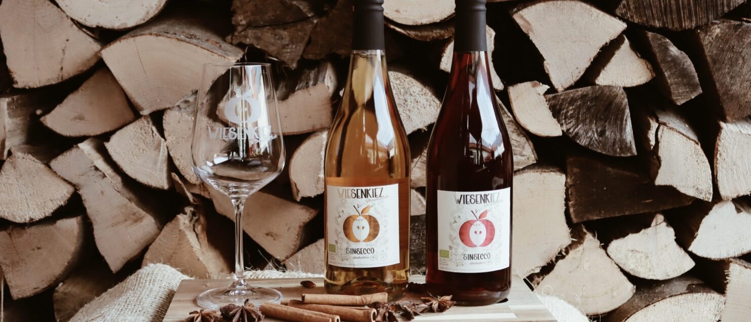 Zwei Flaschen Wiesenkiez SinSecco mit Glas und Zimtstangen vor gestapeltem Kaminholz